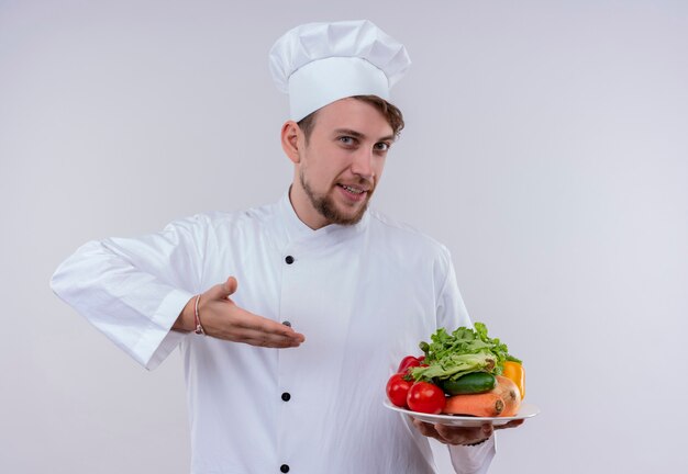 A content young bearded chef man wearing white cooker uniform and hat holding a white plate with fresh vegetables such as tomatoes,cucumbers,lettuce on a white wall