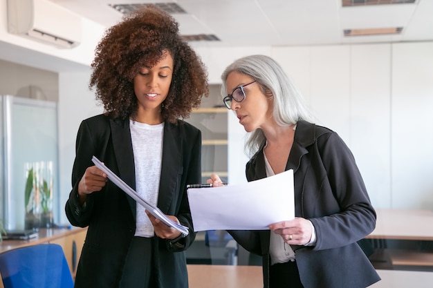 Content young manager showing document to adult colleague. Two pretty content female colleagues holding papers and standing in office room. Teamwork, business and management concept