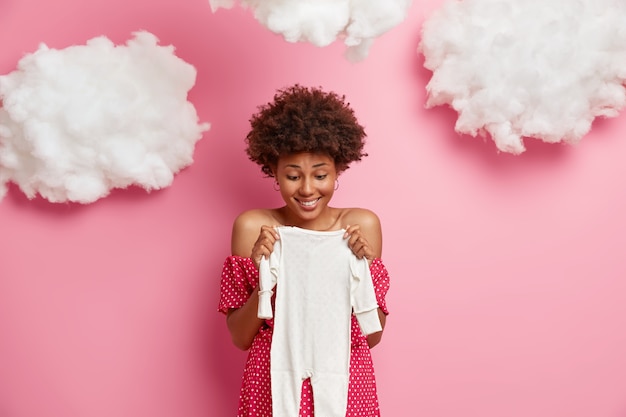 만족스러운 아프리카 계 미국인 임산부, 미래의 아기를위한 바디 수트를 구입하고, 아이의 탄생을 예상하고, 구름 오버 헤드가있는 분홍색 벽에 고립 된, 어린 소녀를 기대합니다. 임신 개념