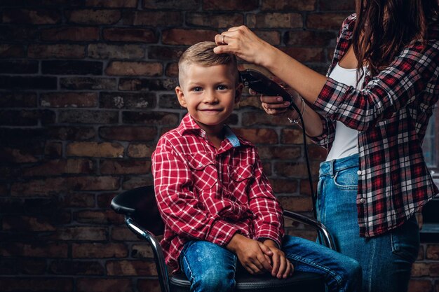 Довольный милый дошкольник стрижется. Детский парикмахер с триммером стрижет маленького мальчика в комнате с лофтовым интерьером.