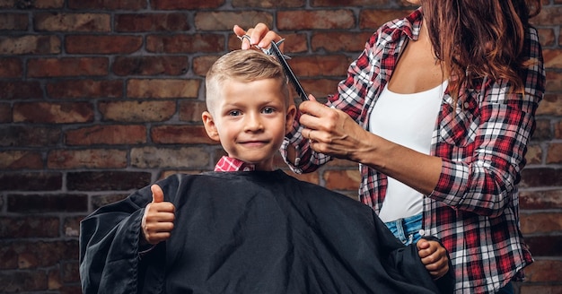 Довольный милый мальчик-дошкольник показывает большой палец вверх во время стрижки. Детский парикмахер с ножницами и расческой стрижет маленького мальчика в комнате с лофтовым интерьером.