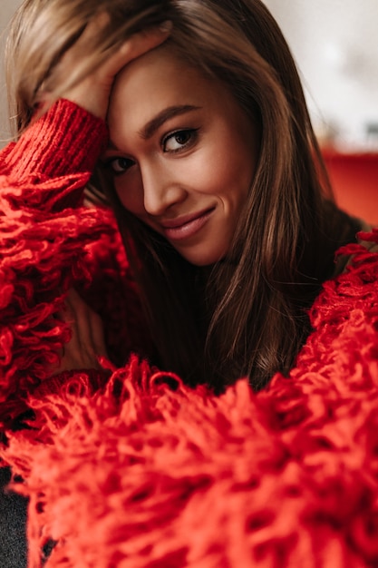 Contented dark-haired woman smiles, leaning over and wrapping herself in red knitted jacket.