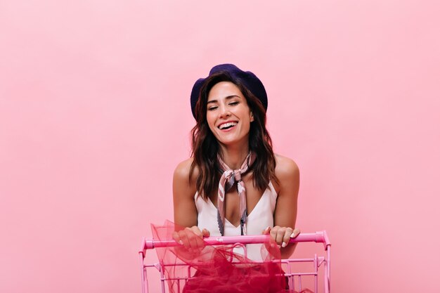 Contented girl sincerely laughs and carries cart on pink background. Smiling woman with wavy dark hair posing and laughing at camera.