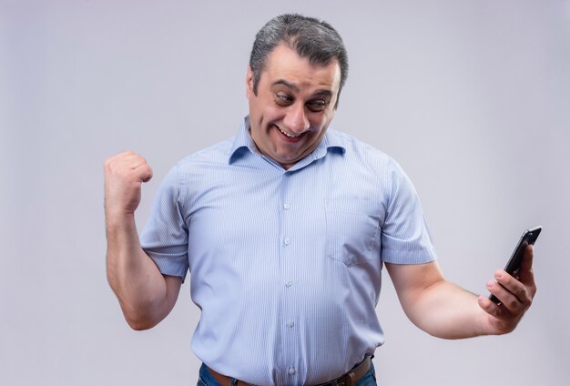 Довольный мужчина средних лет в синей рубашке в вертикальную полоску смотрит в свой мобильный телефон и показывает сжатый кулак жестом руки, стоя