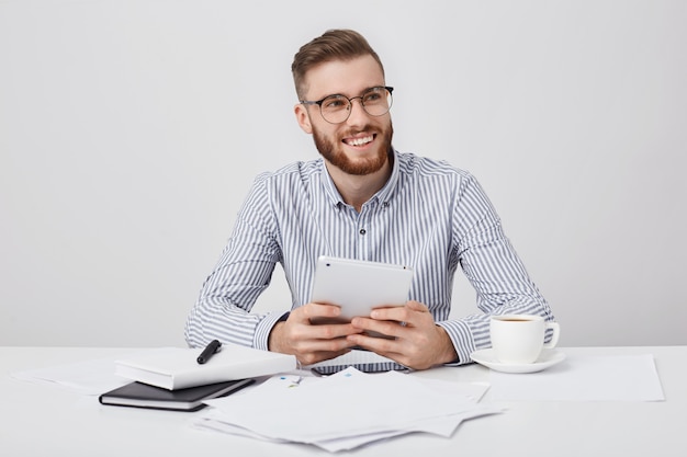 Contented young male freelancer with pleasant smile, holds modern tablet in hands,