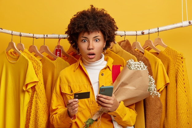 Convenient banking and online shopping concept. Stupefied young African American lady has amazed shocked gaze at camera, holds mobile phone and bouquet, yellow clothes on hangers in background