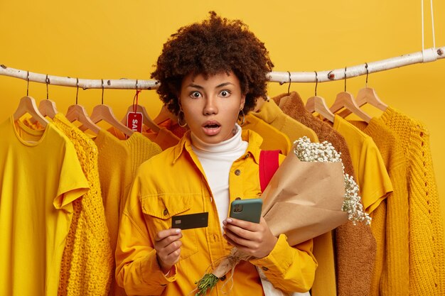 편리한 뱅킹 및 온라인 쇼핑 개념. Stupefied 젊은 아프리카 계 미국인 여성은 카메라에 충격을받은 시선을 놀라게하고 휴대 전화와 꽃다발, 백그라운드에서 옷걸이에 노란색 옷을 보유하고 있습니다.