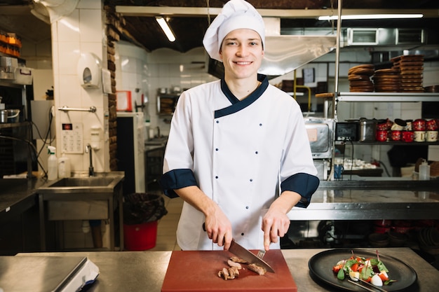 Cook cutting roasted meat on board near salad on plate 