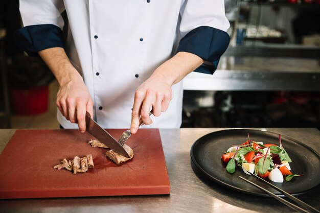 Cook cutting roasted meat on board near salad 