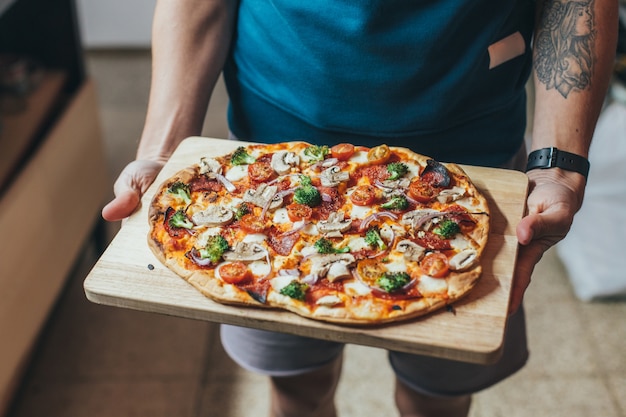 Повар держит деревянный поднос или доску с домашней органической пиццей, покрытой овощами, овощами и сыром.
