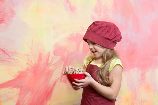 요리사 모자와 앞치마를 입은 요리 아이 또는 어린 소녀는 얼굴에 미소를 띠고 다채로운 배경의 그릇에 쿠키나 비스킷을 들고 공간을 복사합니다.