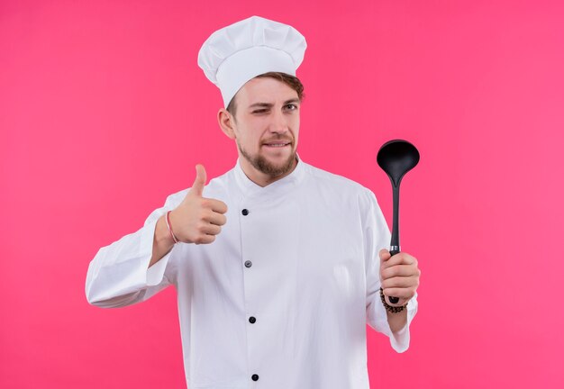 Cook looking at camera shutting one eye showing like gesture with spoon on hand standing over pink wall