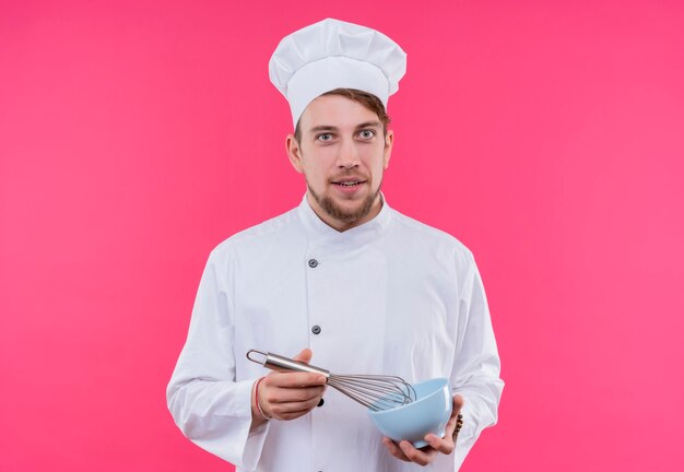 Cook looking at camera smile on face with whisk in bowl standing over pink wall