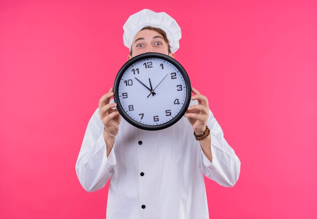 ピンクの壁の上に立っている時計を示す顔にカメラの驚きを見て料理人