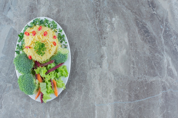 Cooked brown rice served with chopped pepper, cabbage, greens, carrot and broccolie pieces on a platter on marble.