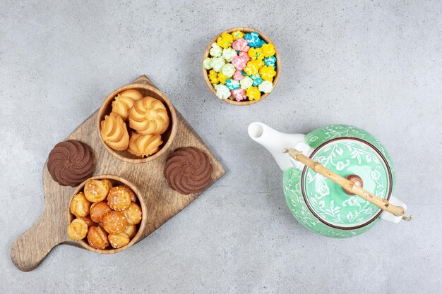 Cookie bowls on wooden board next to ornate teapot and bowl of candies on marble background. High quality photo