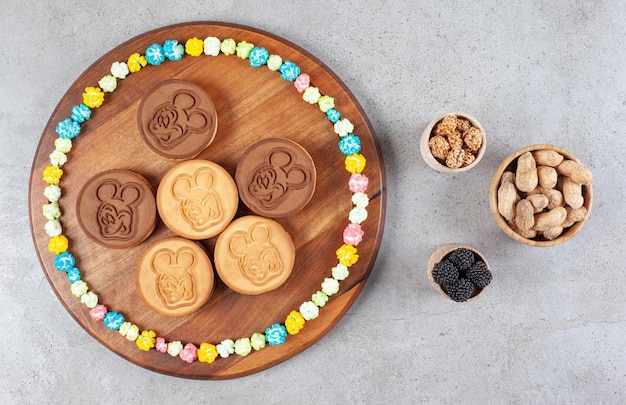 Cookies and a circle of candies on a wooden board next to bowls of peanuts and mullberries on marble background. High quality photo