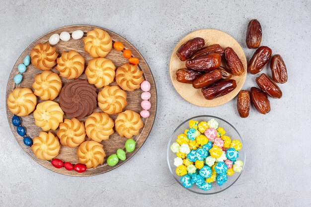 Cookies encircled by candies on wooden board next to bowl of candies and dates on marble surface.