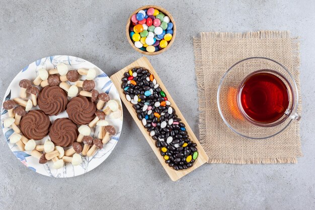 Free photo cookies surrounded by chocolate mushrooms on a plate, candies in a bowl and a tray with a cup of tea on marble surface.