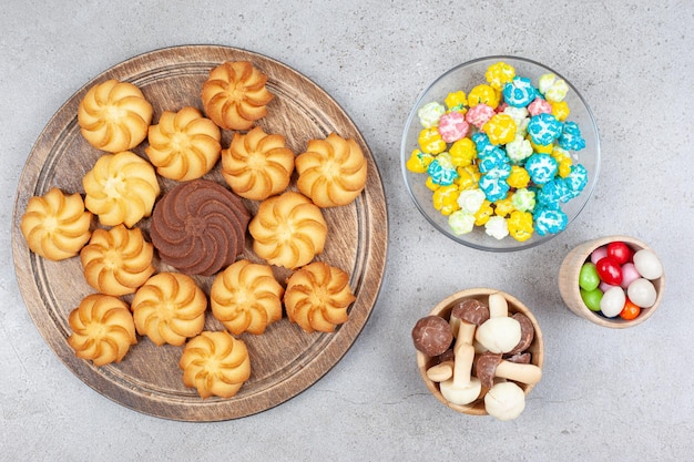 Cookies on wooden board next to bowls of candies and mushroom chocolate on marble surface.