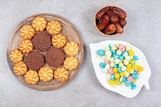 Печенье на деревянной доске рядом с тарелкой конфет и миской фиников на мраморном фоне. Фото высокого качества