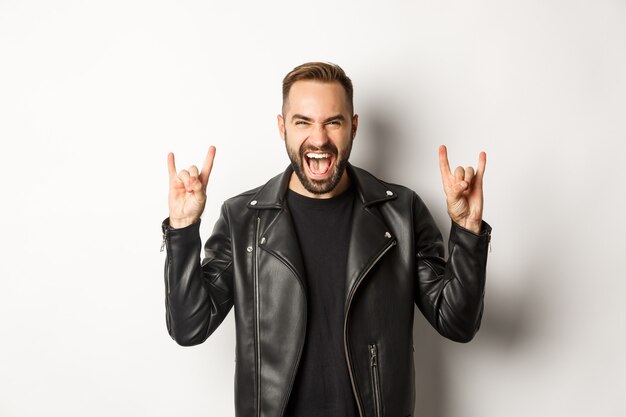 Cool adult man in black leather jacket, showing rock on gesture and tongue, enjoying music festival, standing  