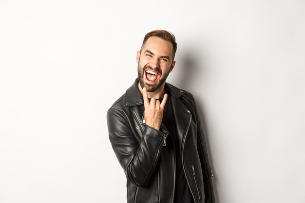 Cool adult man in black leather jacket, showing rock on gesture and tongue, enjoying music festival, standing  