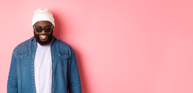 Free photo cool africanamerican hipster man in beanie and sunglasses smiling pleased and looking at camera stan