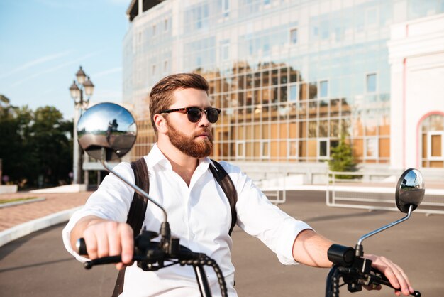 Cool bearded man in sunglasses with backpack rides on modern motorbike outdoors and looking away