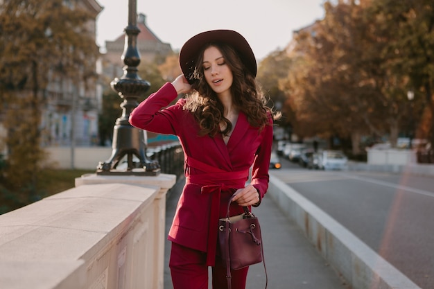 街の通りを歩く紫色のスーツでクールな美しいスタイリッシュな女性、帽子をかぶって、財布を持って春夏秋シーズンのファッショントレンド