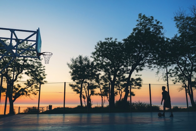 Крутой темнокожий мужчина занимается спортом, играет в баскетбол на восходе солнца, силуэт