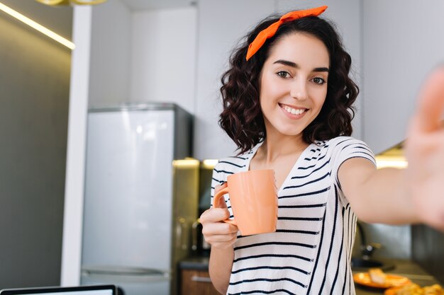 Прохладный яркий портрет селфи привлекательной молодой женщины с вырезанными вьющимися волосами брюнетки, улыбаясь с чашкой чая на кухне современной квартиры. Весело, настоящие положительные эмоции