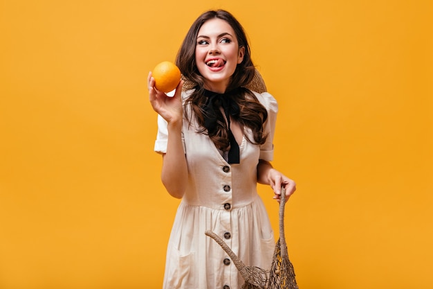 Cool brunette girl in cotton dress with bow around her neck is holding orange and licking.