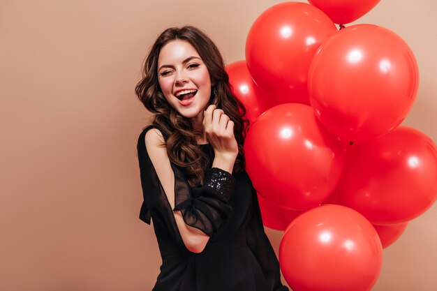 Cool brunette girl in elegant blouse holds bright balloons and winks on isolated background.