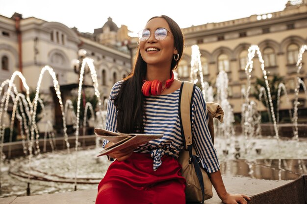 Cool brunette woman in striped shirt, red skirt, headphones and eyeglasses smiles, sits near fountain and holds map