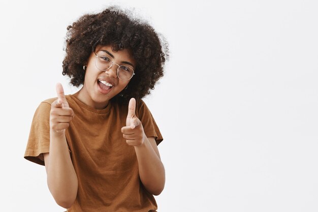 Cool and carefree playful dark-skinned woman in transparent glasses and brown t-shirt tilting head making faces and pointing with finger gun gestures