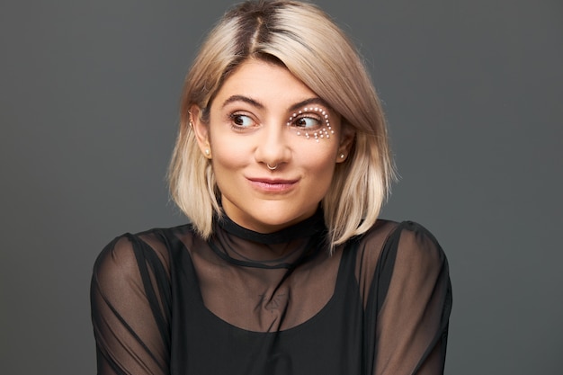 Cool fashionable young caucasian female with stylish haircut and facial piercing thinking about something pleasant, making plans, having great idea, looking away with mysterious enigmatic smile