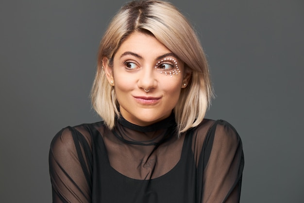 Cool fashionable young Caucasian female with stylish haircut and facial piercing thinking about something pleasant, making plans, having great idea, looking away with mysterious enigmatic smile