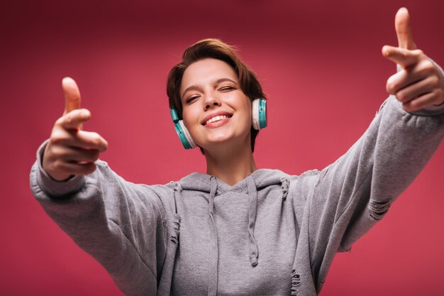 Cool girl in headphones winking and pointing to camera on pink background. Young woman in hoodie listens to music on isolated backdrop