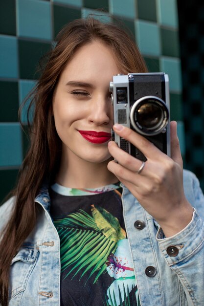 복고풍 필름 카메라를 가진 멋진 소녀 모델은 데님 재킷을 입고, 야외에서 케이지 배경의 도시 벽 위에 검은 머리를 하고 있습니다. 카메라와 함께 도시에서 즐거운 시간을 보내고 사진 작가의 여행 사진.