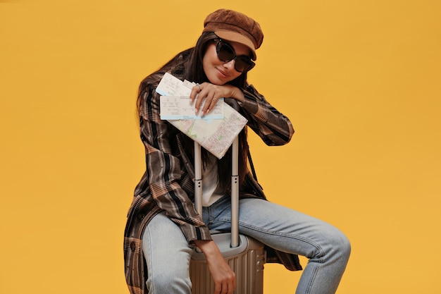Cool girl in sunglasses beret and stylish coat smiles softly and sits on suitcase on isolated Brunette woman in denim pants poses and holds map on yellow background