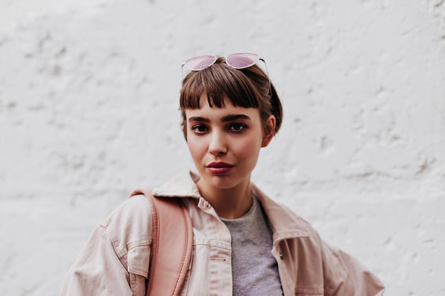 Cool girl with short hair looking into camera on background on white backdrop Brunette lady with glass in beige outside posing on backdrop of wall