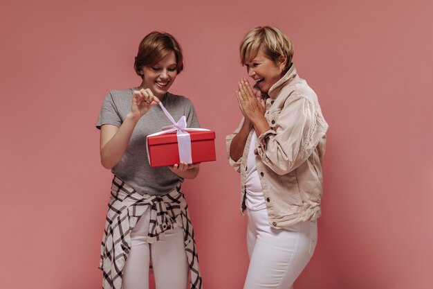 Cool girl with short hairstyle in t-shirt, plaid shirt and light pants opening red gift box and posing with cheerful old woman on pink backdrop. 