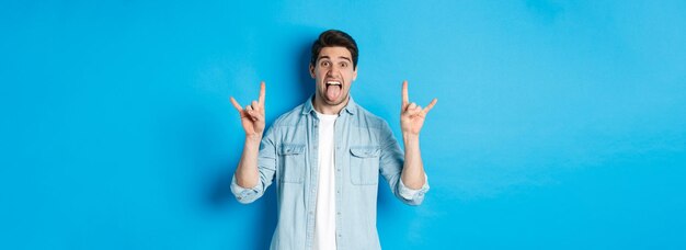 Cool guy having fun enjoying concert showing rock sign and tongue standing over blue background