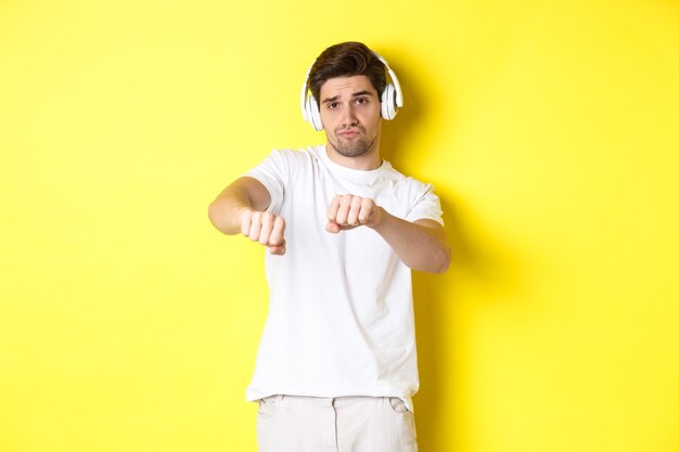Крутой парень слушает музыку в наушниках и танцует, стоя в белой одежде на желтом фоне студии.