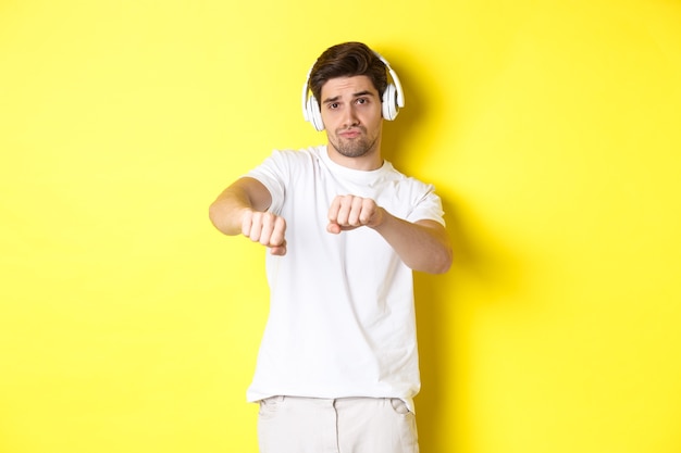 Cool guy listening music in headphones and dancing, standing in white clothes against yellow studio background.