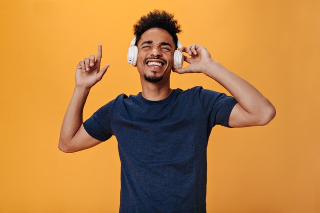 Крутой парень показывает палец вверх и слушает музыку на оранжевом фоне Портрет темнокожего мужчины в синей футболке, танцующего на изолированном фоне
