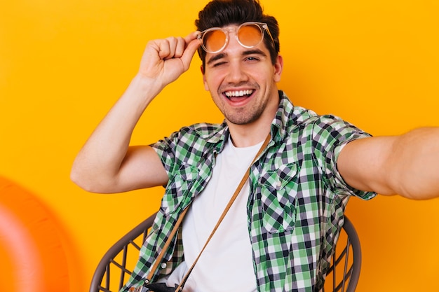 Free photo cool guy in white t-shirt and plaid shirt takes off his glasses, winks and takes selfie on orange space.