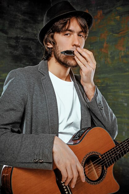회색 스튜디오 배경에 기타와 하모니카를 연주하는 모자와 함께 멋진 남자