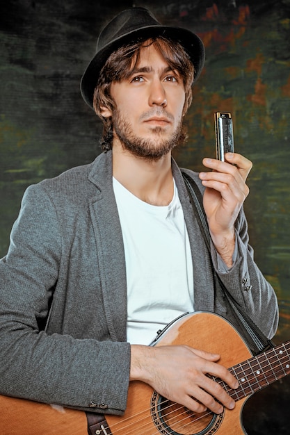 Крутой парень в шляпе играет на гитаре и губной гармошке на сером студийном фоне